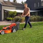 Husqvarna batteridriven gräsklippare LC142i utan batteri & laddare fotograferad från sidan medan en man går bakom och klipper gräset med gräsklipparen med ett svart hus i bakgrunden